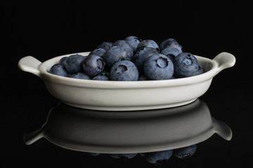 Lot of whole fresh blue bilberry in white oval ceramic bowl isolated on black glass