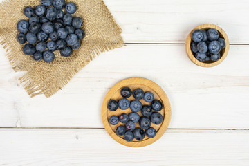 Lot of whole fresh blue bilberry in tiny wooden bowl on round bamboo coaster on natural sackcloth flatlay on white wood