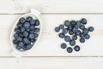 Lot of whole fresh blue bilberry in white oval ceramic bowl flatlay on white wood