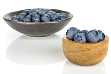 Lot of whole fresh blue bilberry in tiny wooden bowl in dark ceramic bowl isolated on white background
