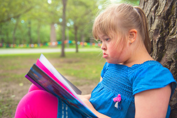 A portrait of trisomie 21 child girl outside having fun on a park reading book