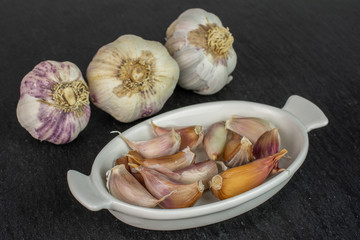 Group of three whole lot of pieces of organic white garlic allium sativum in white oval ceramic bowl on grey stone