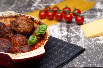 meatballs with tomato sauce and decorated with basil leaf, served in white and red pan on grey background. next to the pan near fresh tomatoes, spaghetti and a piece of hard cheese. Closeup.
