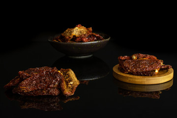 Lot of whole dried red tomato in dark ceramic bowl on round bamboo coaster isolated on black glass