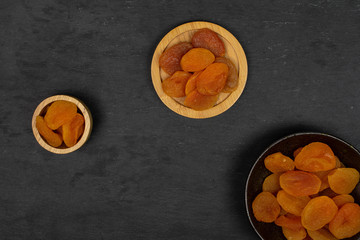 Lot of whole dried orange apricot in tiny wooden bowl on round bamboo coaster in dark ceramic bowl flatlay on grey stone