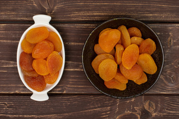 Lot of whole dried orange apricot in white oval ceramic bowl in dark ceramic bowl flatlay on brown wood