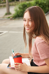 school girl writing pen in notebook on the knees. Student with coffee in a paper cup near university.