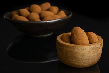 Lot of whole sweet brown chocolate cinnamon almond in tiny wooden bowl in dark ceramic bowl isolated on black glass