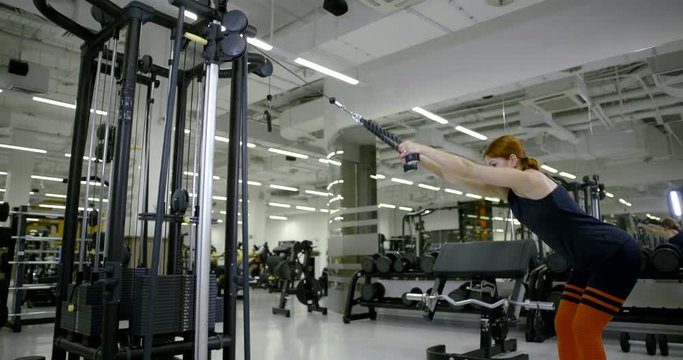 A slender red-haired woman coach is engaged in a block simulator, shakes her back. She is in the gym.