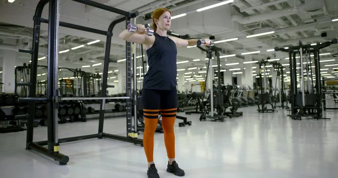 Athletic girl in the gym doing standing exercises with dumbbells on her arms and shoulders.
