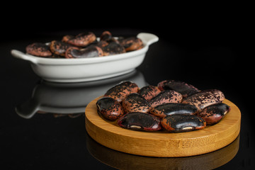 Lot of whole stained purple bean on round bamboo coaster in white oval ceramic bowl isolated on black glass