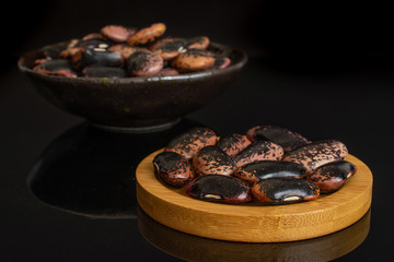 Lot of whole stained purple bean on round bamboo coaster in dark ceramic bowl isolated on black glass