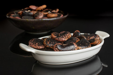 Lot of whole stained purple bean in white oval ceramic bowl in dark ceramic bowl isolated on black glass