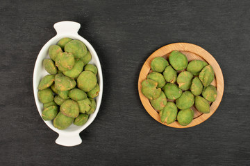Lot of whole spicy green wasabi peanut in white oval ceramic bowl on bamboo coaster flatlay on grey stone