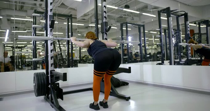 Sports girl in the gym doing standing exercises with a barbell on his back, raises and lowers the shoulders of the bar, shoulders and back, bends the ass back. Rear view.