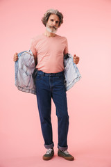 Naklejka premium Full length portrait of stylish mature man 70s with gray beard looking at camera and holding his denim jacket