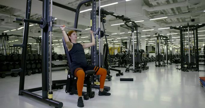 Sports girl in the gym doing sitting exercises on the shoulders, press with a barbell, raises and lowers the barbell, shakes his hands and shoulders.