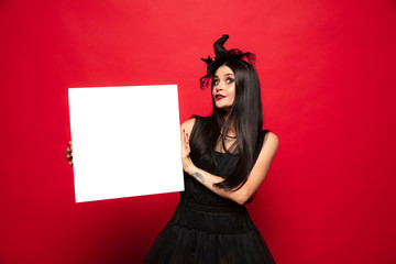 Young brunette woman in black hat and costume on red background. Attractive caucasian female model. Halloween, black friday, cyber monday, sales, autumn concept. Holding empty sheet for copyspace.