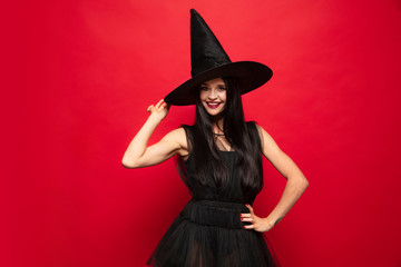 Young brunette woman in black hat and costume on red background. Attractive caucasian female model. Halloween, black friday, cyber monday, sales, autumn concept. Copyspace. Dancing, posing charming.