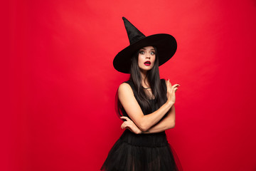 Young brunette woman in black hat and costume on red background. Attractive caucasian female model. Halloween, black friday, cyber monday, sales, autumn concept. Copyspace. Pointing up.