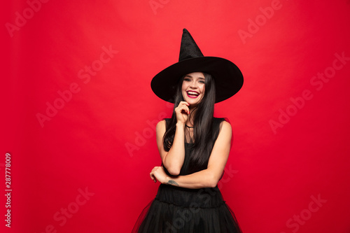 Young brunette woman in black hat and costume on red background. Attractive caucasian female model. Halloween, black friday, cyber monday, sales, autumn concept. Happy smiling.