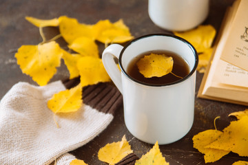 two white cups with tea in yellow leaves with a book and a knitted scarf, the concept of warmth and autumn sadness