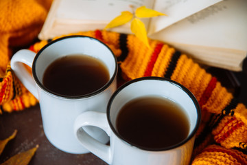 mugs surrounded by yellow foliage with a book and a knitted scarf, the concept of warmth and autumn sadness