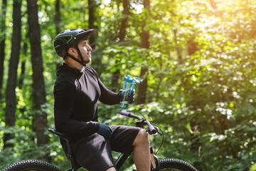 Male cyclist sitting on bike and drinking water in woods
