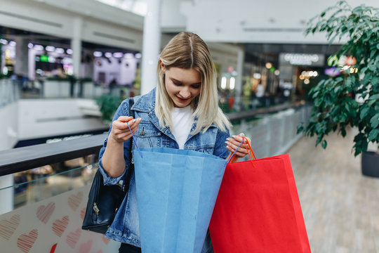 Commerce and shopping season. Beautiful girl in a denim jacket with bags on blurred shopping mall background. Woman looking in her shopping bag. Copy space on the left or right side