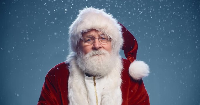 Senior man in santa clause outfit looking at camera, smiling and rubbing his hands, isolated on blue snowy background - christmas spirit concept close up 4k footage