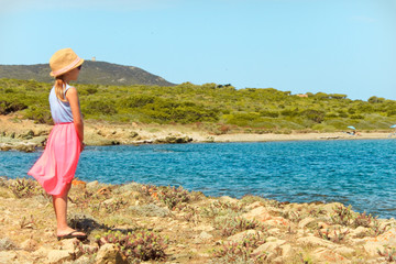 Petite fille en tenue rose et bleu avec chapeau de paille devant la mer bleue turquoise regardant au loin