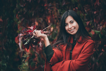 Joyful beautiful girl with long natural black hair on autumn background of green red grape hedge. Happy girl in red shawl and green coat with grape bouquet. Female emotional portrait in faded tones.