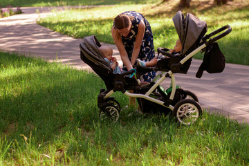 Woman and stroller for twins. A beautiful woman in a park seats two children up to a year in a...