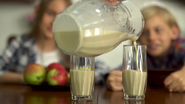 Female hand pouring milk from a jug in the glasses for the kids in the foreground. Two children fool around in the background. Healthy lifestyle. Focus in the foreground.