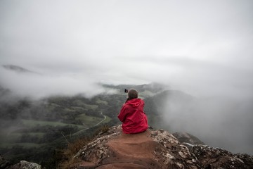 Garota de vermelho fazendo selfie no alto de uma montanha entre as nuvens