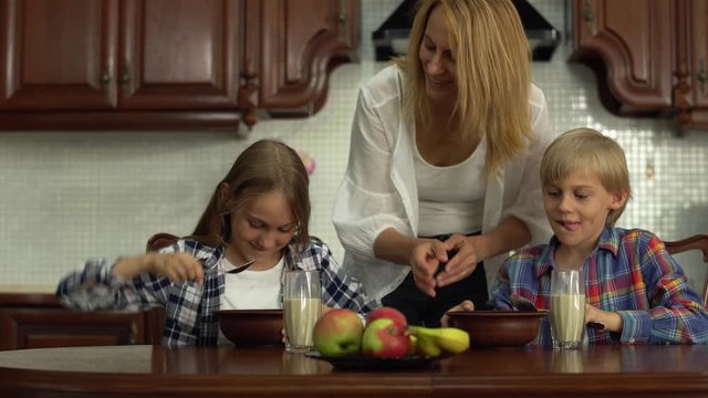 Happy mother feeding kids before school in the morning. Smiling siblings eating cereals and drinking milk at the table in the kitchen. The woman kissing kids. Happy friendly family enjoying breakfast