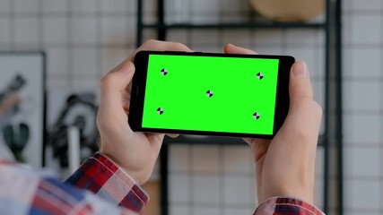 Man hands holding black smartphone mobile device with blank green display in home interior - over shoulder close up view. Mock up, copyspace, chroma key, template, green screen, technology concept