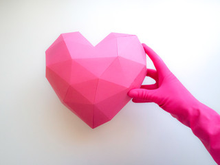 A pink gloved hand holding a pink paper heart of multiple faces, crafted in low poly technique, over a white background