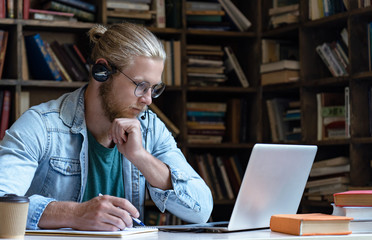 Focused serious young man wear wireless headset look at laptop screen make notes study e learning...