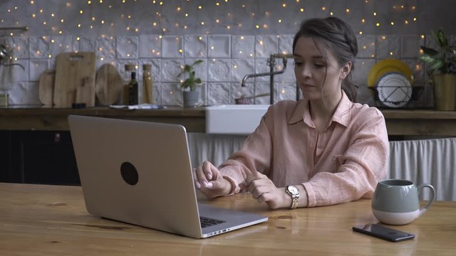 Lovely young woman sitting in the kitchen at wooden table and typing on her laptop with the cup of tea or coffee. Stock footage. Modern decorated home interior on the background