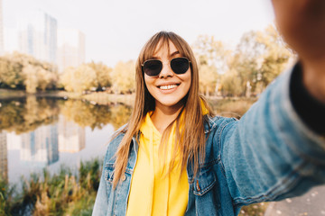 Autumn walk. Woman portrait. Happy girl in jean jacket and sun glasses is looking at camera and smiling while walking in the park near the lake