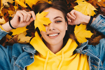 Autumn walk. Woman portrait. Happy girl in yellow hoodie and jean jacket is playing with leaf, looking at camera and smiling while lying on the ground in the park; top view