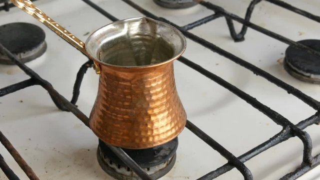 A teaspoon puts black ground coffee in a copper Turk which stands on a white gas stove