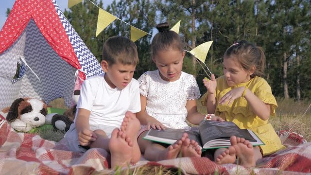 interaction of children, cute little females and a charismatic male child are reading book for children with interest looking at pictures of relaxing in forest on the background of a wigwam