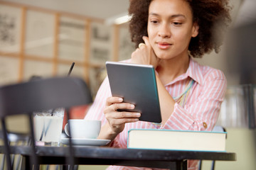 Fototapeta premium Attractive mixed race woman in striped pink dress sitting in cafe and using tablet.