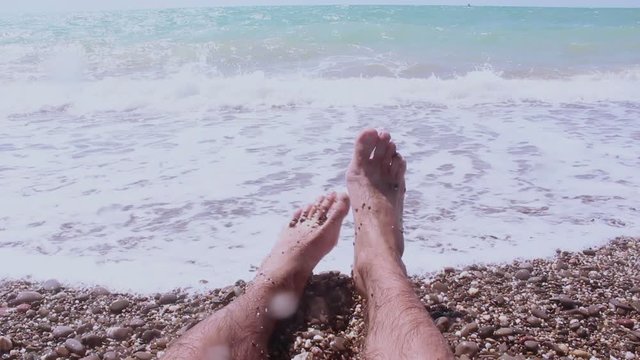 View of the legs of a man lying on the seashore. A man enjoys a vacation on a pebble beach. Male legs on a background of blue sea.