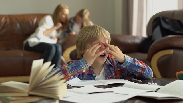 Tired little boy sitting at the table with notebooks at home. Mother and sister sitting in the background looking pappers. Adorable schoolkids doing exercise for tired eyes while doing lessons.