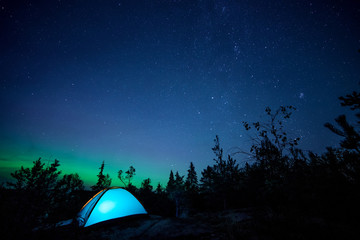 Night scene with illuminated camping tent, forest, starry sky and northern lights