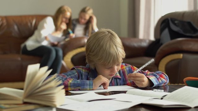 Little boy sitting at the table with notebooks at home while doing homework. Mother and sister sitting in the background looking pappers. Schoolboy rubs tired eyes with his hand.