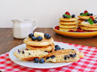 Blueberry mini pancakes with syrup on white plate  over red and white checkered, gingham napkin. Stack of silver dollar pancakes.